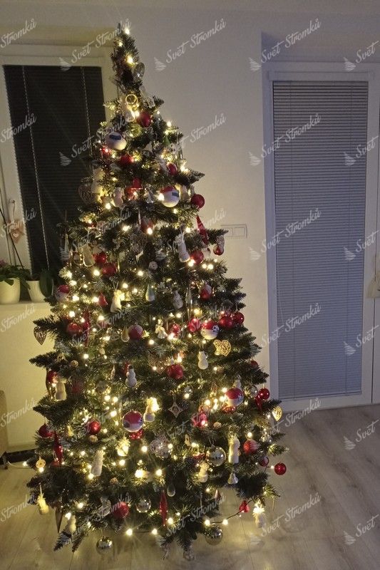 Klasický umělý vánoční stromeček s tmavě zelenými větvičkami, stříbrné konce větviček s krystaly ledu, hustě ozdobený červeno-bílými ozdobami a teplým bílým osvětlením v obýváku